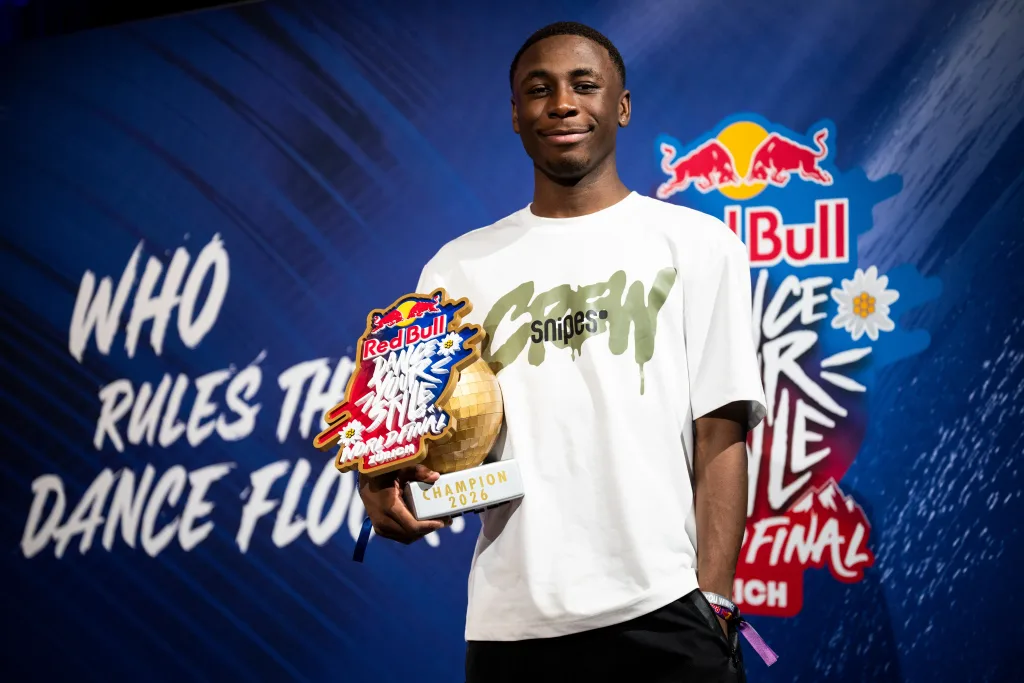 Willis, in a white T-shirt and smiling, holds the gold disco-ball "Champion 2026" Red Bull Dance Your Style trophy in front of the official event backdrop
